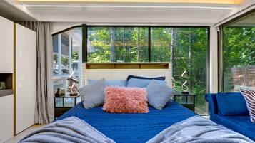 A modern bedroom with a large bed, a view of trees through large windows, and a blue sofa.