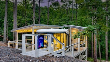 A modern cabin with a glass enclosure, surrounded by a deck, set in a forested area.