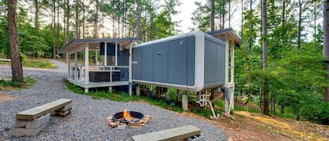 Une cabane avec un foyer extérieur, une terrasse couverte et une grande boîte grise contenant des équipements.