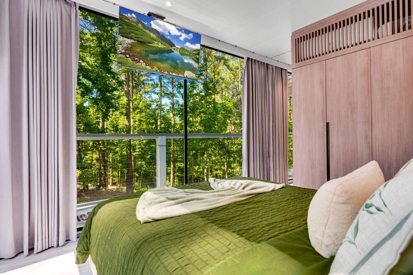A bedroom with a large window, a bed with a green bedspread, and a view of a forest.