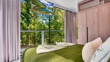 A bedroom with a large window, a bed with a green bedspread, and a view of a forest.