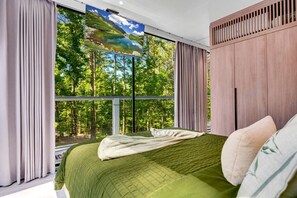 Un dormitorio con un ventanal grande, una cama con edredón verde y vistas a un bosque.