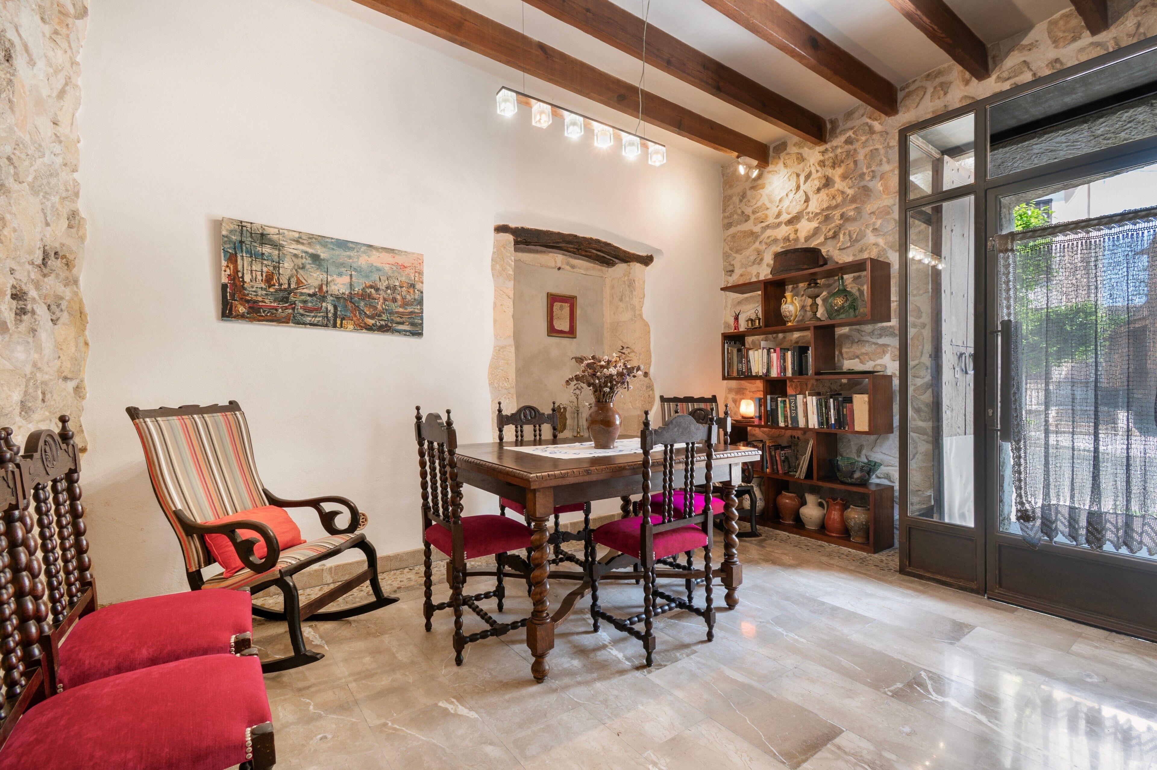 A dining area with a wooden table, chairs, a bookshelf, and a stone wall.