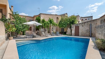 A swimming pool with lounge chairs and umbrellas, surrounded by stone buildings with arched windows.