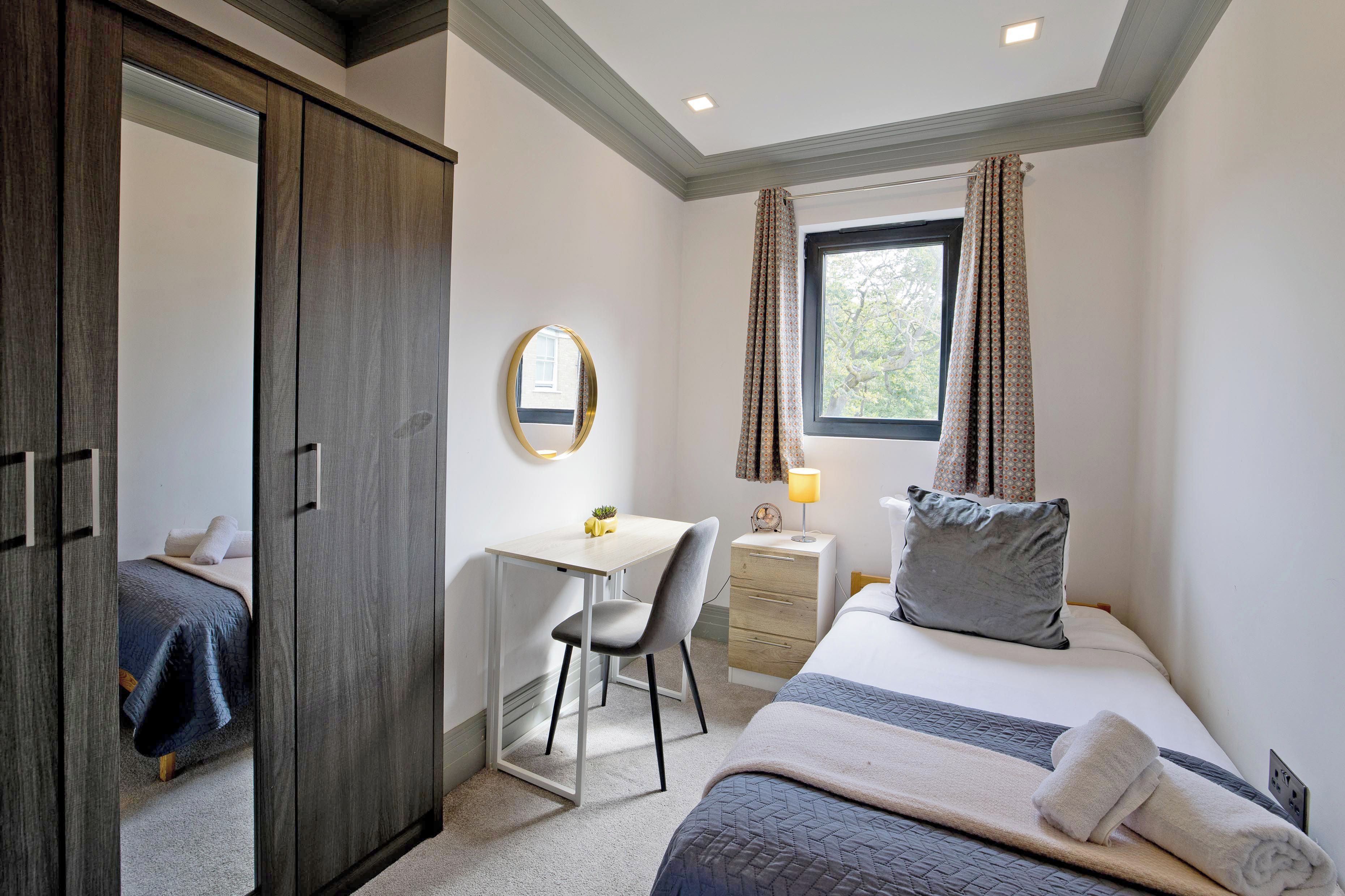 A bedroom with a bed, a desk, a chair, a wardrobe, a mirror, and a window with curtains.
