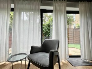 A room with a single grey chair, a small round table, and a view of a backyard through large windows with white curtains. - Luxury 4BR Camden Villa w/ Parking & Garden (London)