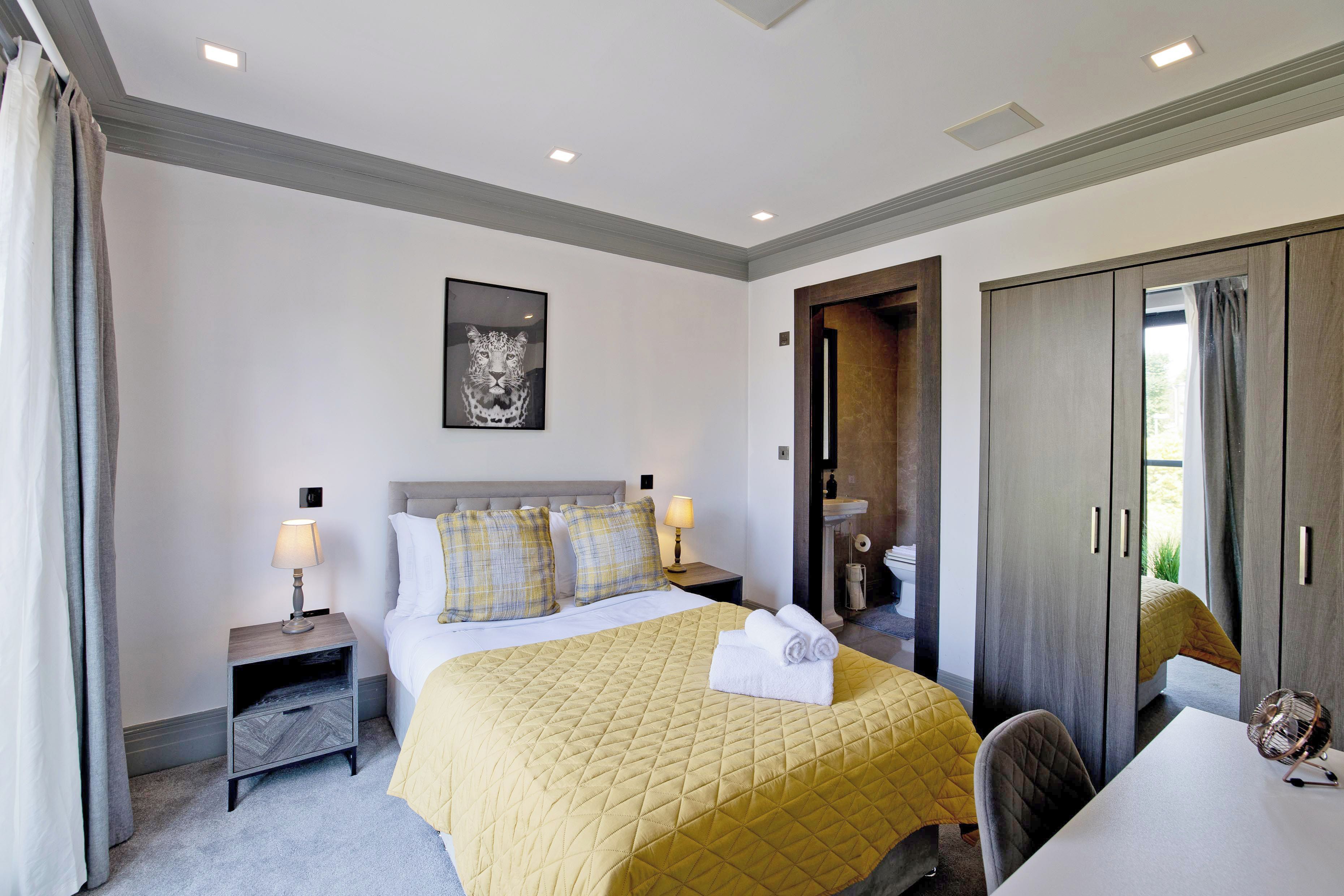 A hotel room with a bed, bedside tables, a desk, a chair, a wardrobe, and a bathroom visible through an open door.