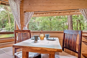 A wooden table with chairs, a pitcher, and a mug on a balcony with curtains and a view of trees. - Treehouse 'Wernerwald' with Balcony and Wi-Fi (Cuxhaven)