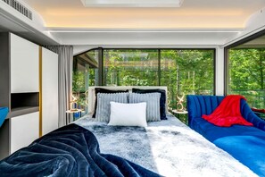 Ein modernes Schlafzimmer mit einem großen Bett, einem blauen Sofa und Blick auf Grün durch große Fenster.