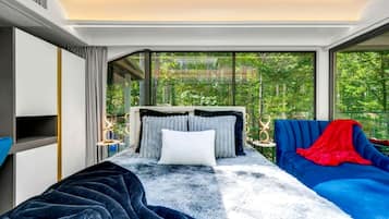 A modern bedroom with a large bed, a blue sofa, and a view of greenery through large windows.