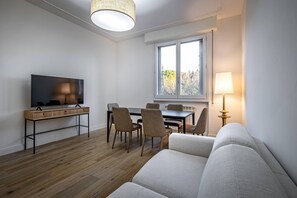 A modern living room with a sofa, a TV on a wooden stand, a dining table with chairs, and a window with a view of trees. - Pietrafitta (Florence)