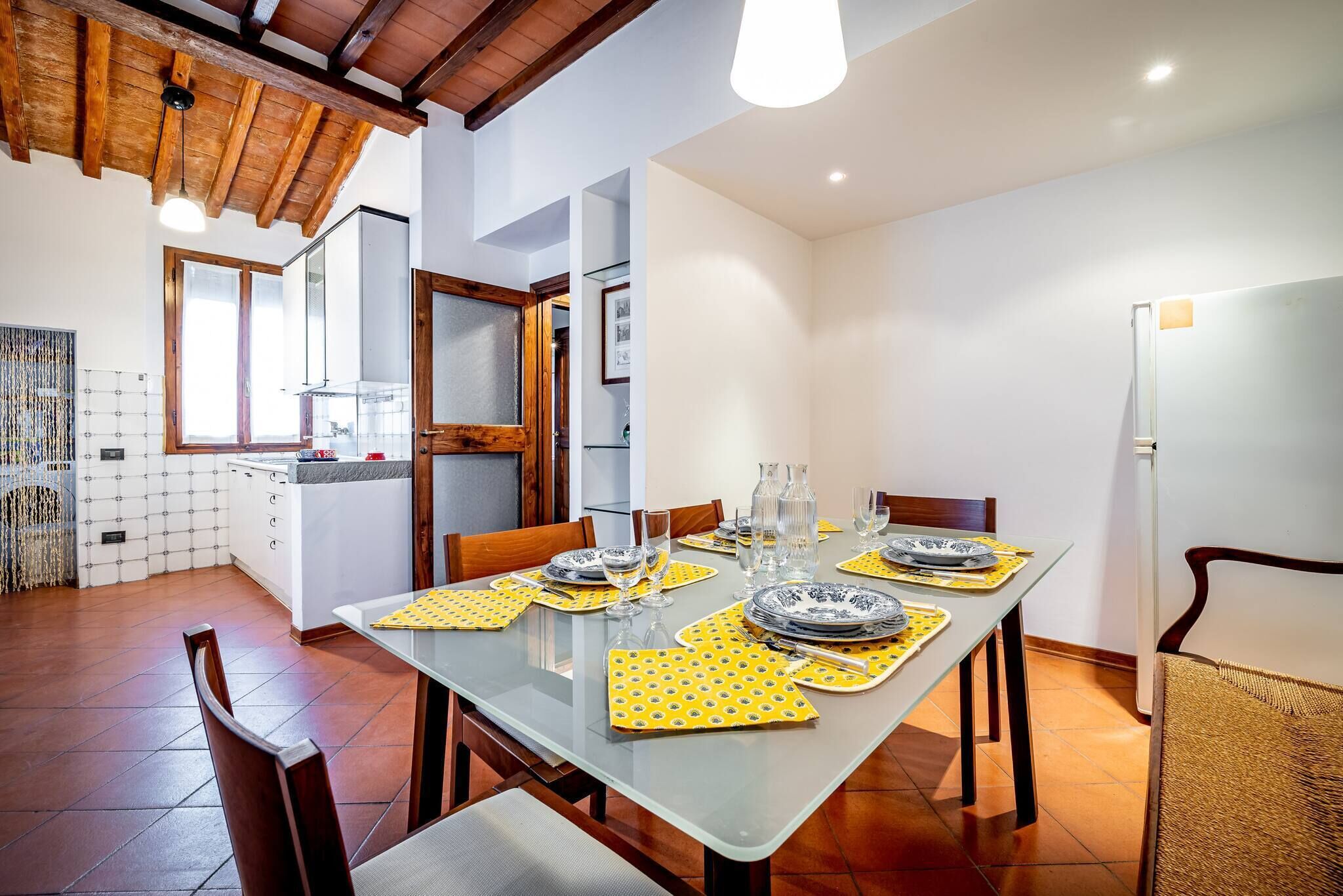 A dining area with a glass table set for four, wooden chairs, and a kitchen in the background.