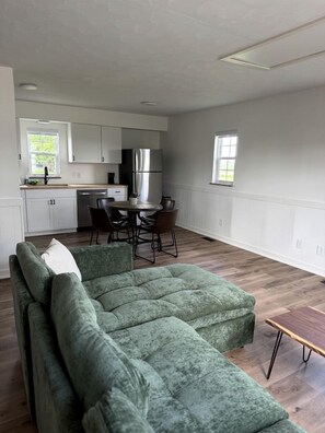 Ett modernt vardagsrum med en hörnsoffa, ett matbord med stolar, en köksdel med skåp och vitvaror samt ett fönster som släpper in naturligt ljus.