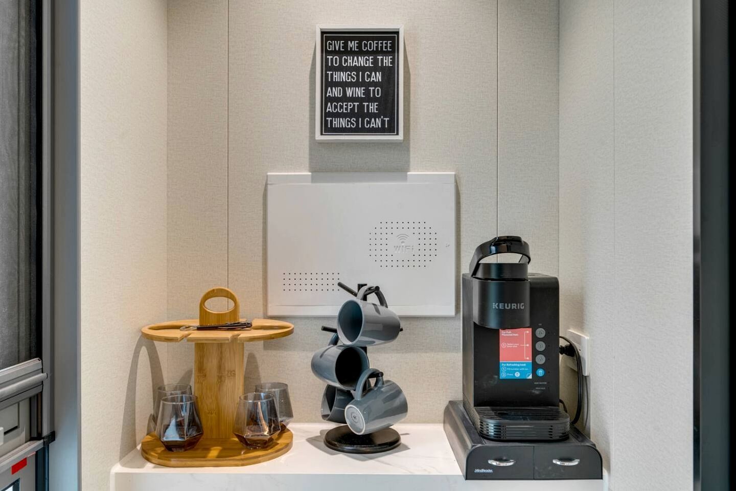 Una estación de café con una máquina Keurig, un letrero, una bandeja de madera con vasos y un panel en la pared.