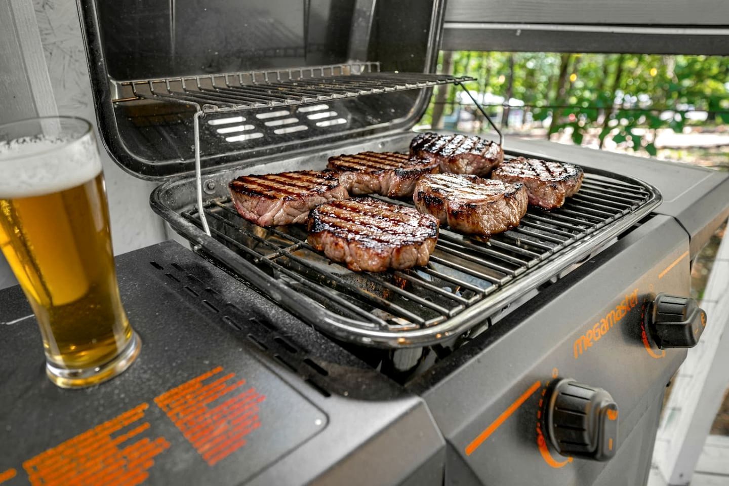 A grill with several steaks cooking, a glass of beer, and a grill with instructions.