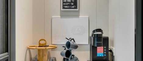 Una estación de café con una máquina Keurig, un letrero, una bandeja de madera con vasos y un panel en la pared.