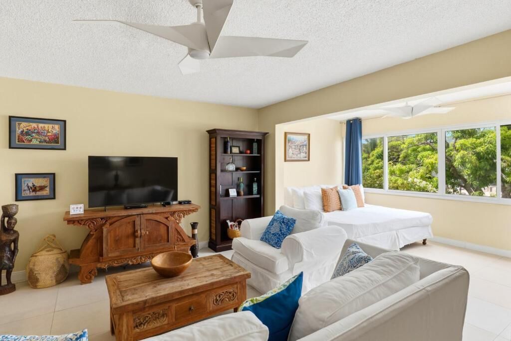 A spacious living room with a television, wooden furniture, a sofa, a bed, and a large window with a view of trees.