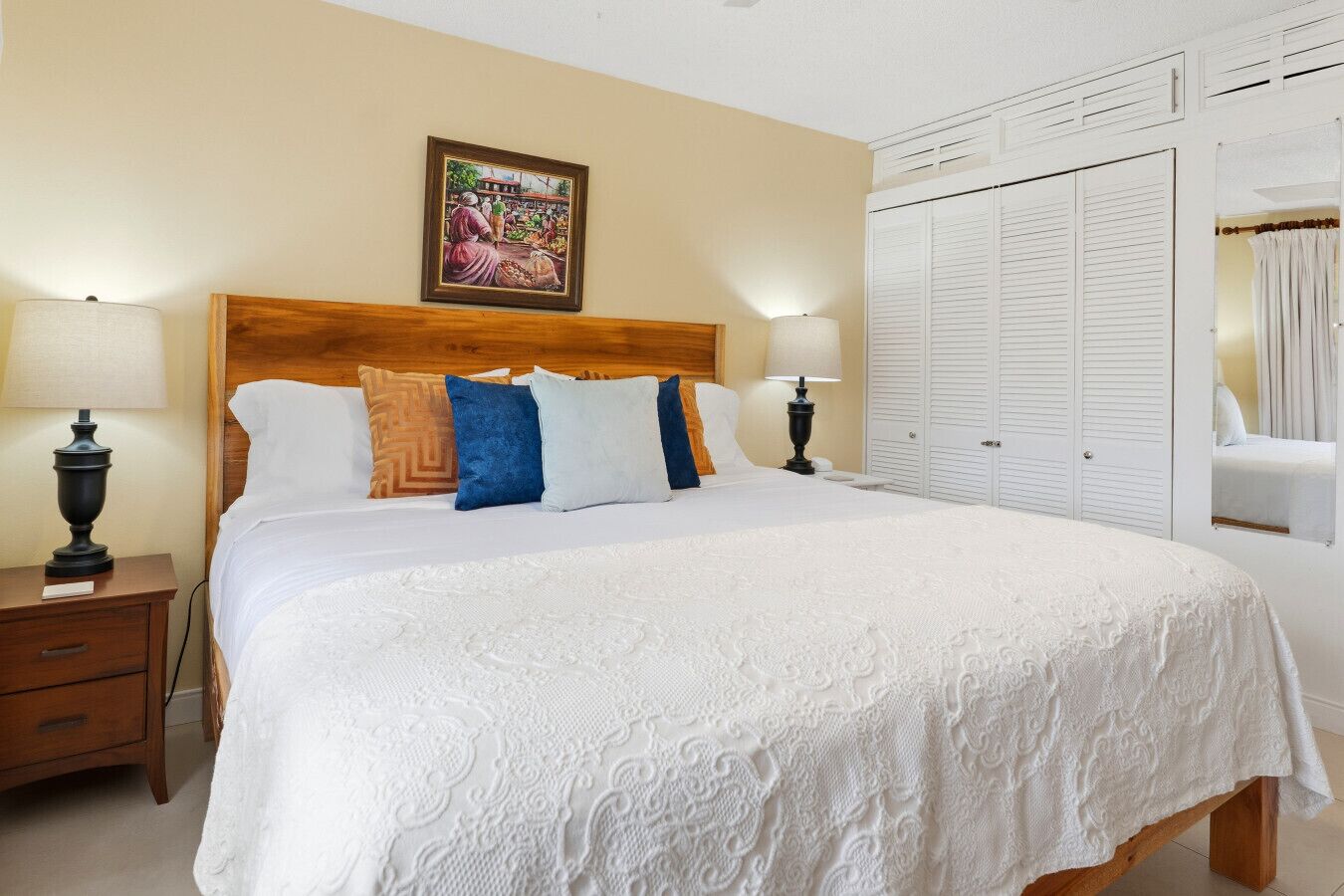 A bedroom with a wooden headboard, white bedding, a nightstand with a lamp, a wardrobe with louvered doors, and a framed picture on the wall.