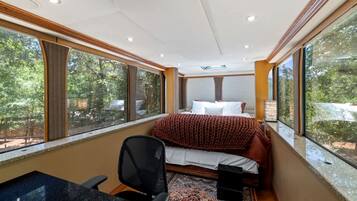 A bedroom with a bed, a desk, and a chair. There are large windows on one side.