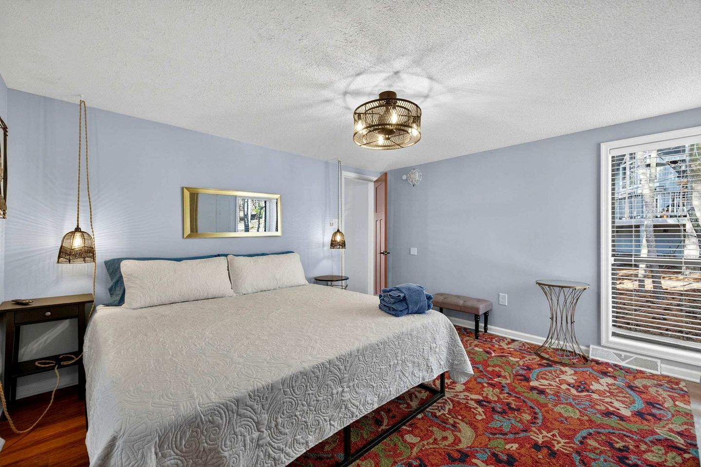 A bedroom with a large bed, a mirror, a hanging light fixture, a bench, and a view of the city through the window.