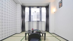 A room with tatami flooring, a low table with a cushion, a small table with a vase, and a window with curtains. - Spacious 4-bedroom apartment in heart of Osaka with AC (Osaka)