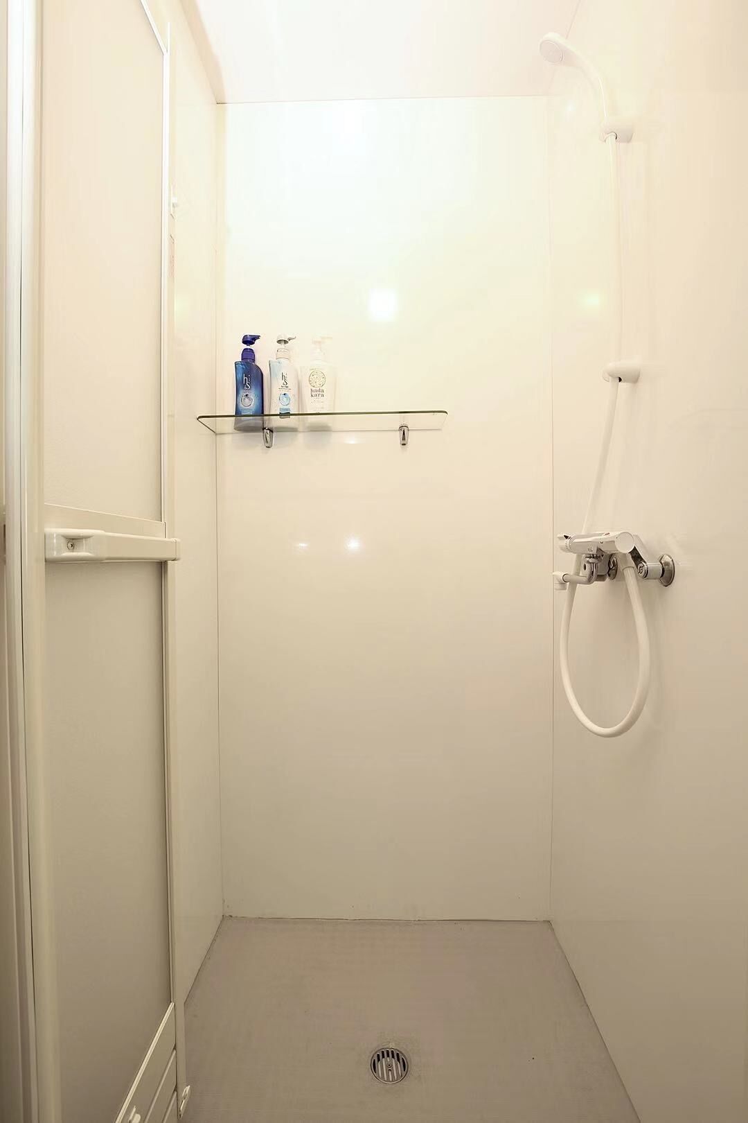 A small, white bathroom with a shower, a glass shelf with toiletries, and a door.