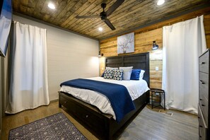 A bedroom with a bed, a ceiling fan, a nightstand, a dresser, and a wall-mounted lamp. - Luxury Forest Cabin with Hot Tub, Fire Pit & Game Room Near Broken Bow Lake, Broken Bow, Oklahoma (Broken Bow)