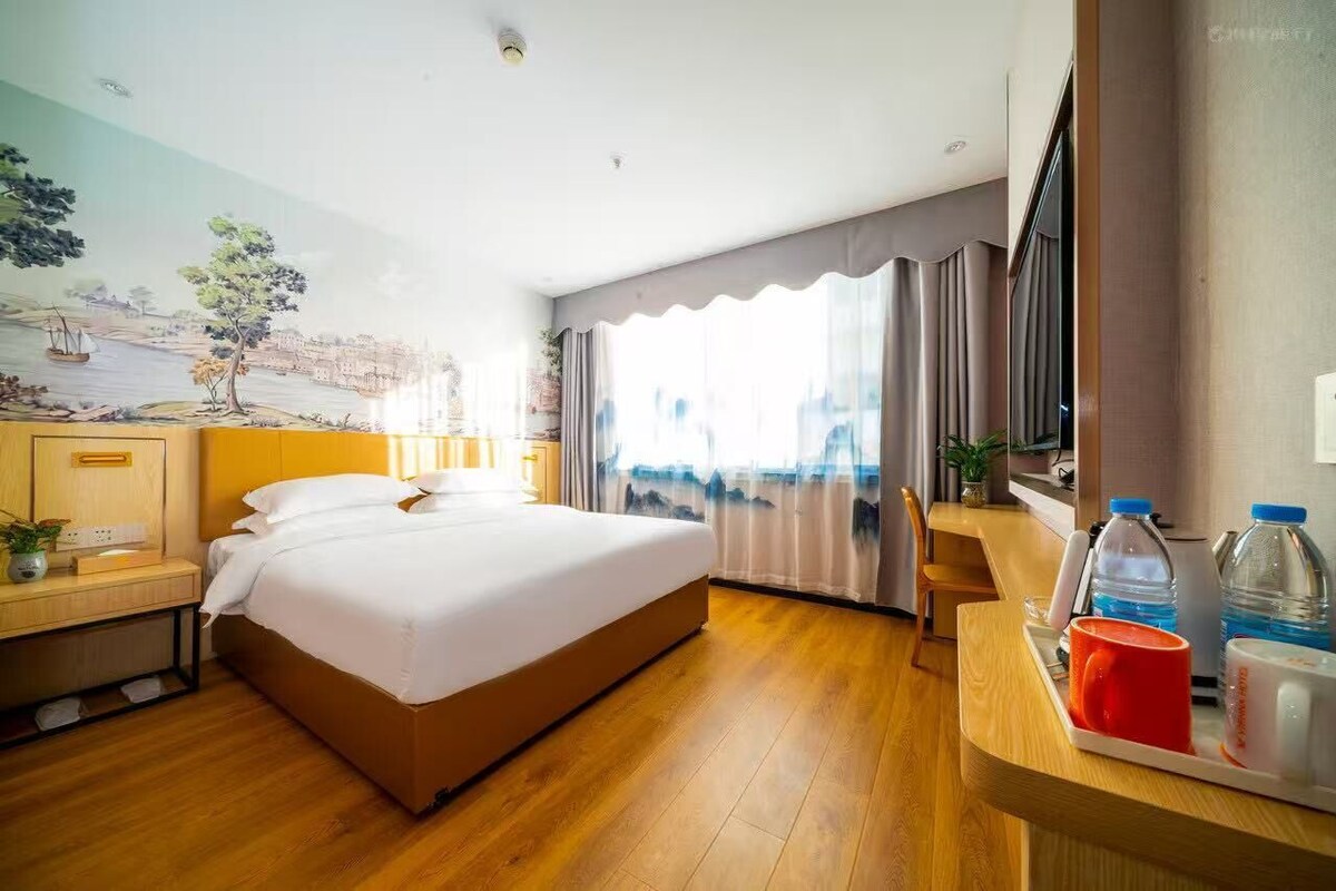 A hotel room with a large bed, a desk with a chair, a window with curtains, and a mural on the wall.