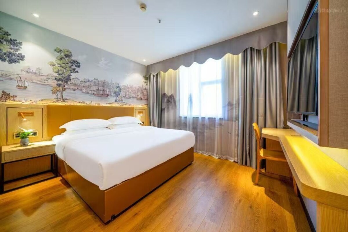 A hotel room with a bed, a desk, a chair, a nightstand, a wall mural, and a window with curtains.