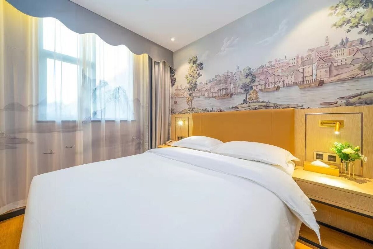 A hotel room with a bed, a nightstand, a wall mural, and a window with curtains.