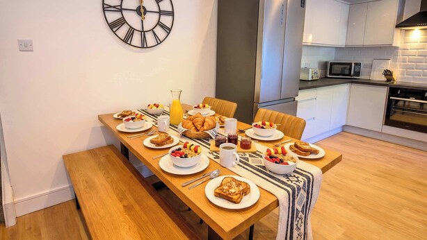 Uma área de refeições com mesa de madeira posta para o café da manhã, um relógio na parede e uma cozinha moderna ao fundo.