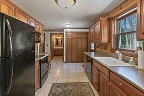 A kitchen with wooden cabinets, a black refrigerator, a sink, and a window with a view of trees. - Cozy Forest Cabin with Fireplace Near Lake Michigan Beaches in Manistee, Michigan (Manistee)