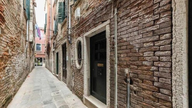 A narrow alleyway with brick buildings, a stone-paved path, and a black door on the right. - Cà Turchette (Venezia)