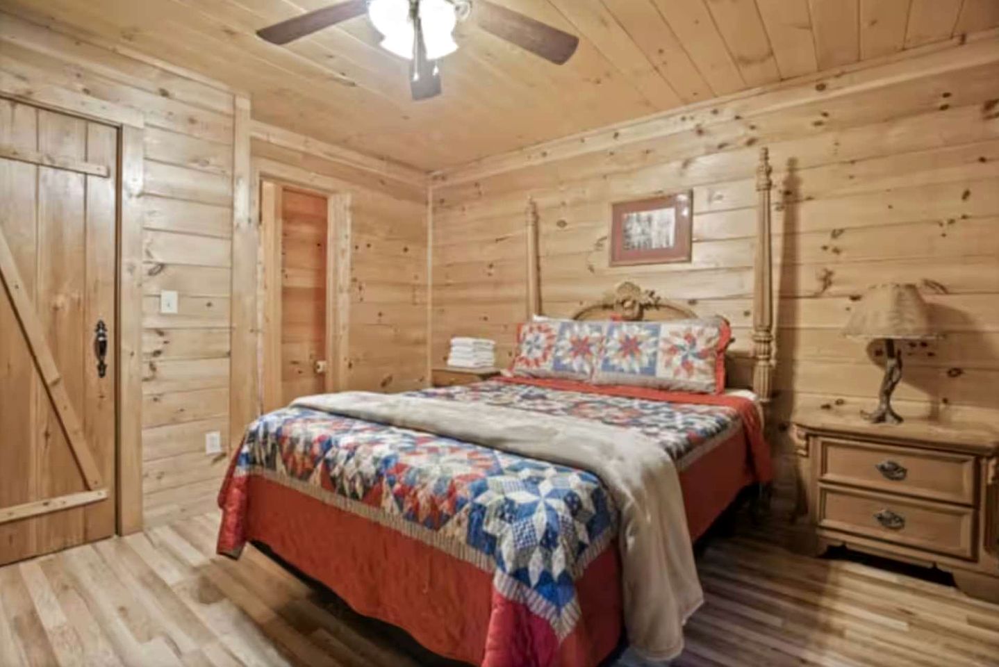 Un dormitorio de cabaña de madera con cama, mesita de noche, cómoda, ventilador de techo y puerta.