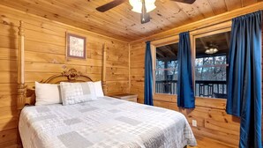 Een slaapkamer in een houten chalet, met een bed, plafondventilator, raam met gordijnen en een lijst met foto.