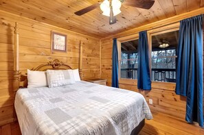 Un dormitorio de cabaña de madera con una cama, un ventilador de techo, una ventana con cortinas y un cuadro enmarcado en la pared.
