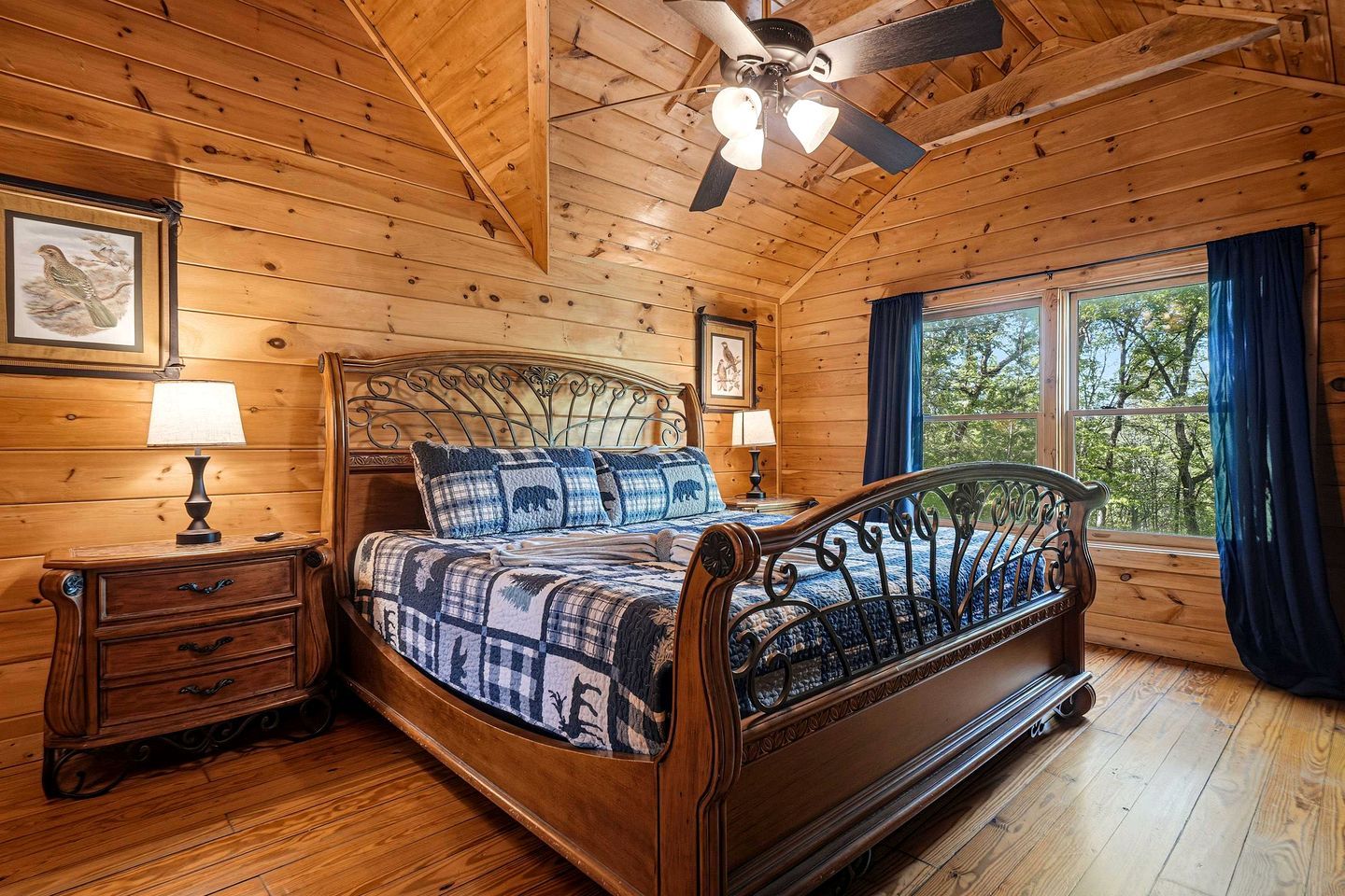 A wooden cabin bedroom with a bed, nightstands, a ceiling fan, and a window with curtains.
