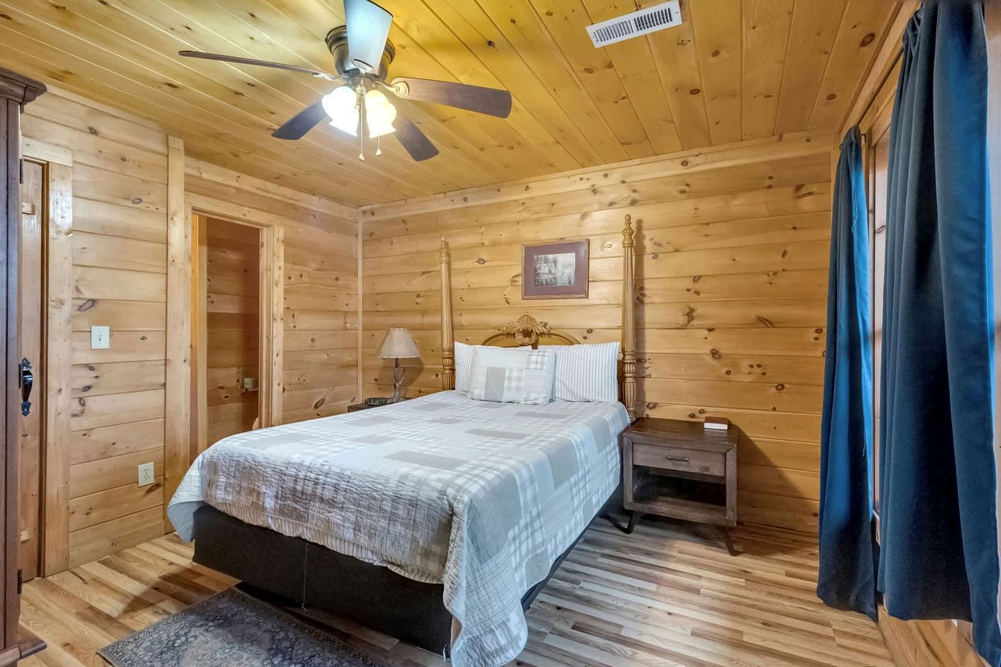 Un dormitorio con paneles de madera, una cama, una mesita de noche, una lámpara, ventilador de techo y una puerta.