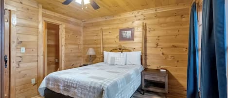 A wooden-paneled bedroom with a bed, nightstand, lamp, ceiling fan, and a door.