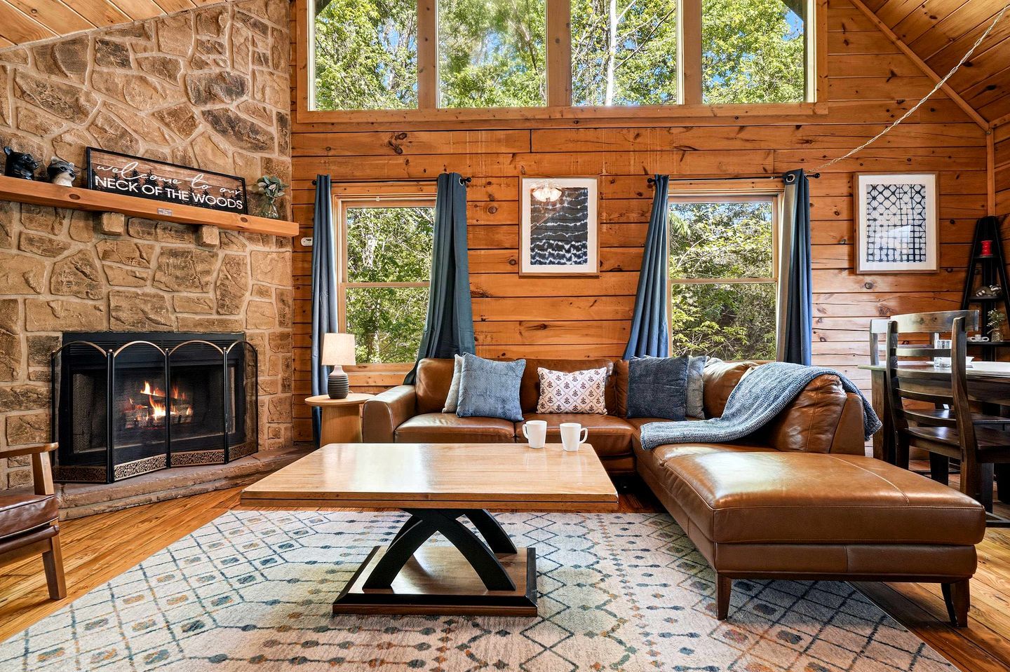 A cozy cabin interior with a stone fireplace, leather furniture, and large windows.