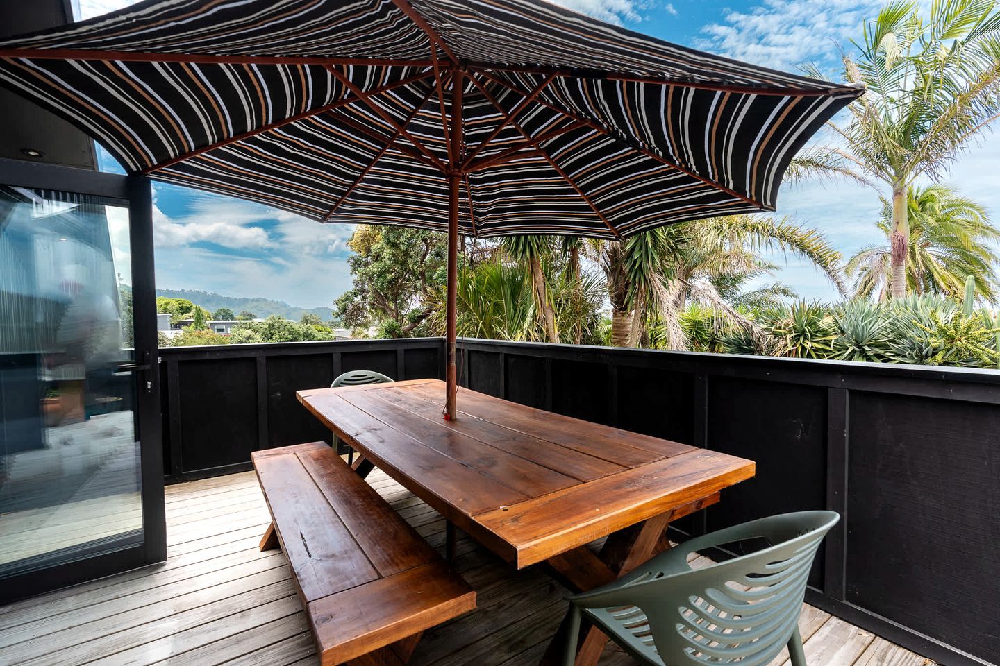 A wooden deck with a picnic table and chairs, an umbrella, and a view of palm trees and mountains.