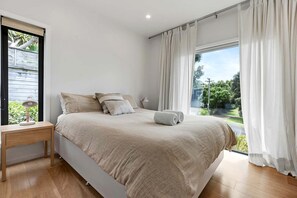 Een slaapkamer met een bed, nachtkastje, lamp en uitzicht op groen.