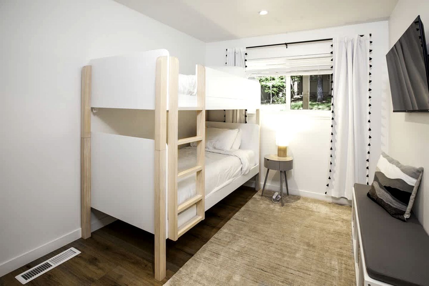 Ein Stockbett mit einer Leiter, ein Nachttisch mit Lampe, ein Fenster mit Vorhängen und ein an der Wand befestigter Fernseher.