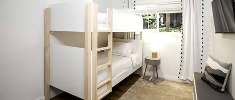 A bunk bed with a ladder, a bedside table with a lamp, a window with curtains, and a TV mounted on the wall.