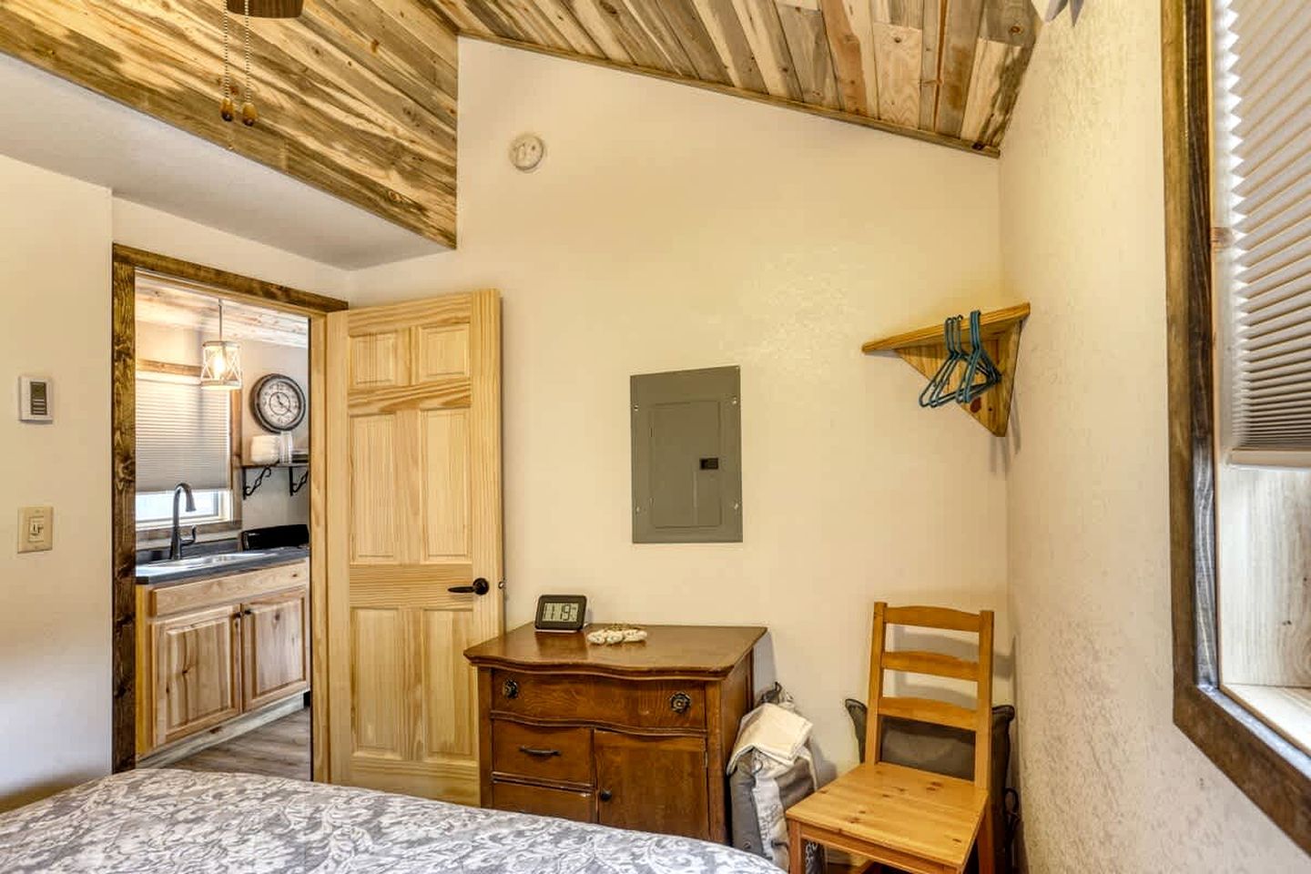 Ein gemütlicher Raum mit Bett, Holzkommode, kleinem Tisch, Stuhl, einer Uhr und einem Fenster mit Jalousien.