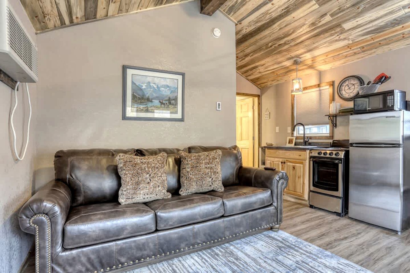 A living room with a leather sofa, a framed picture on the wall, a kitchen with a stove and refrigerator, and a wooden ceiling.