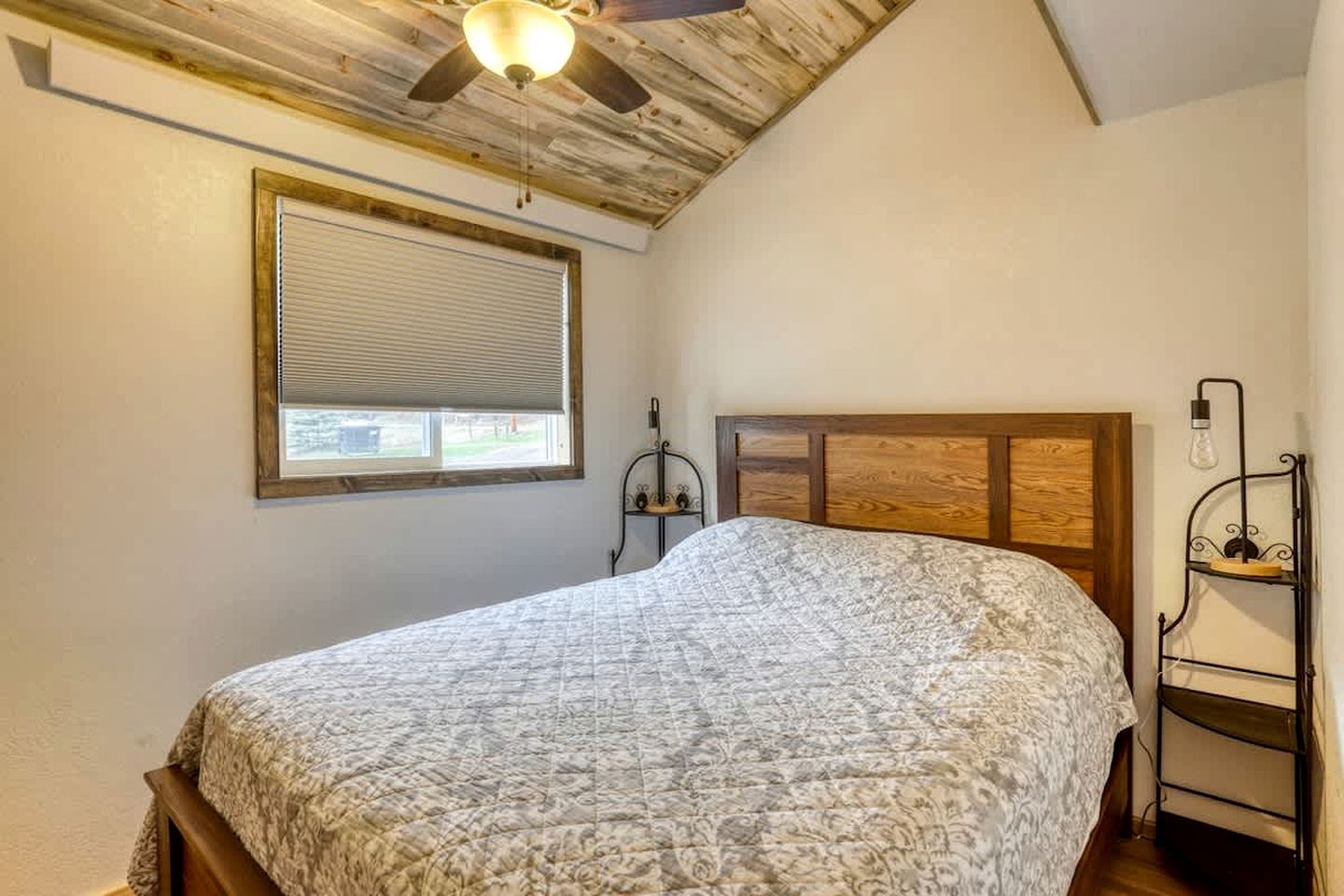 Ein Schlafzimmer mit Holzboden, einem Bett mit gemustertem Überwurf, einem Fenster mit Jalousien und einem Metallregal mit Glühbirne.