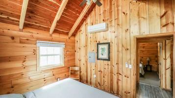 A wooden cabin room with a bed, a ceiling fan, a window, and a framed picture on the wall.
