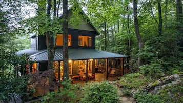 A two-story cabin with a covered porch, surrounded by dense forest.