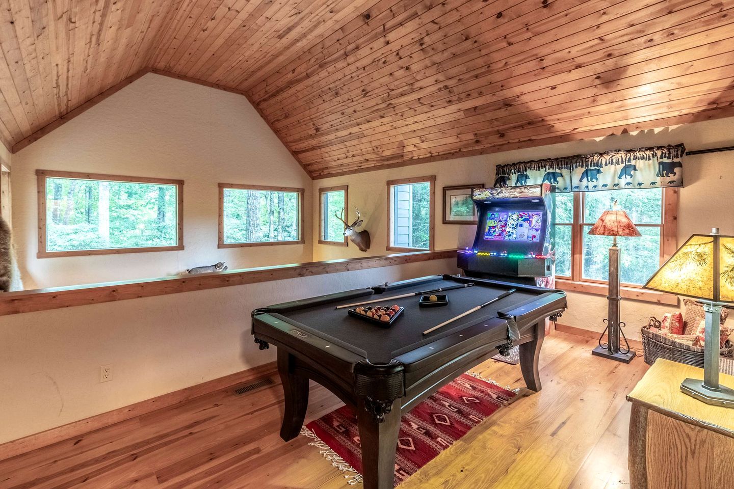 A pool table with a ceiling fan, a mounted deer head, and a video game console in a room with wooden walls and windows.